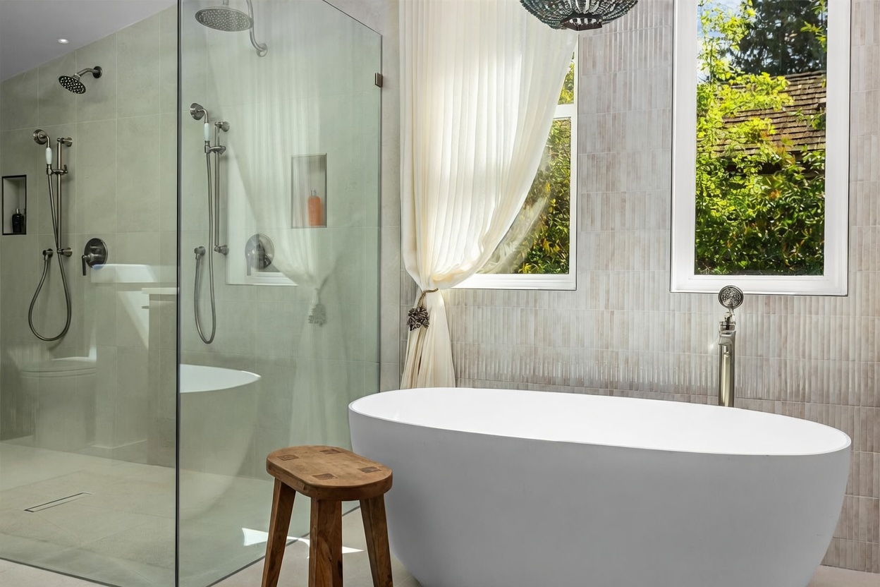 This elegant primary bathroom features a sleek, freestanding soaking tub positioned next to a large window with soft, draped curtains. To the left, a spacious walk-in shower is enclosed by glass panels and includes both a rainfall showerhead and a handheld sprayer. A small, rustic wooden stool sits in the foreground, adding a warm, organic touch to the neutral-toned, tiled space.
