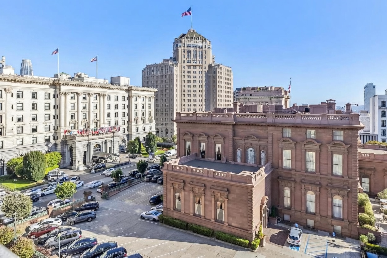 This high-angle aerial view captures a historic, multi-story brownstone building with classical architectural details, situated in a dense urban environment. The scene includes a paved parking lot in the foreground and a prominent, ornate hotel building with flags in the background, set against a clear blue sky. The perspective provides a comprehensive look at the property's location within a bustling city neighborhood.