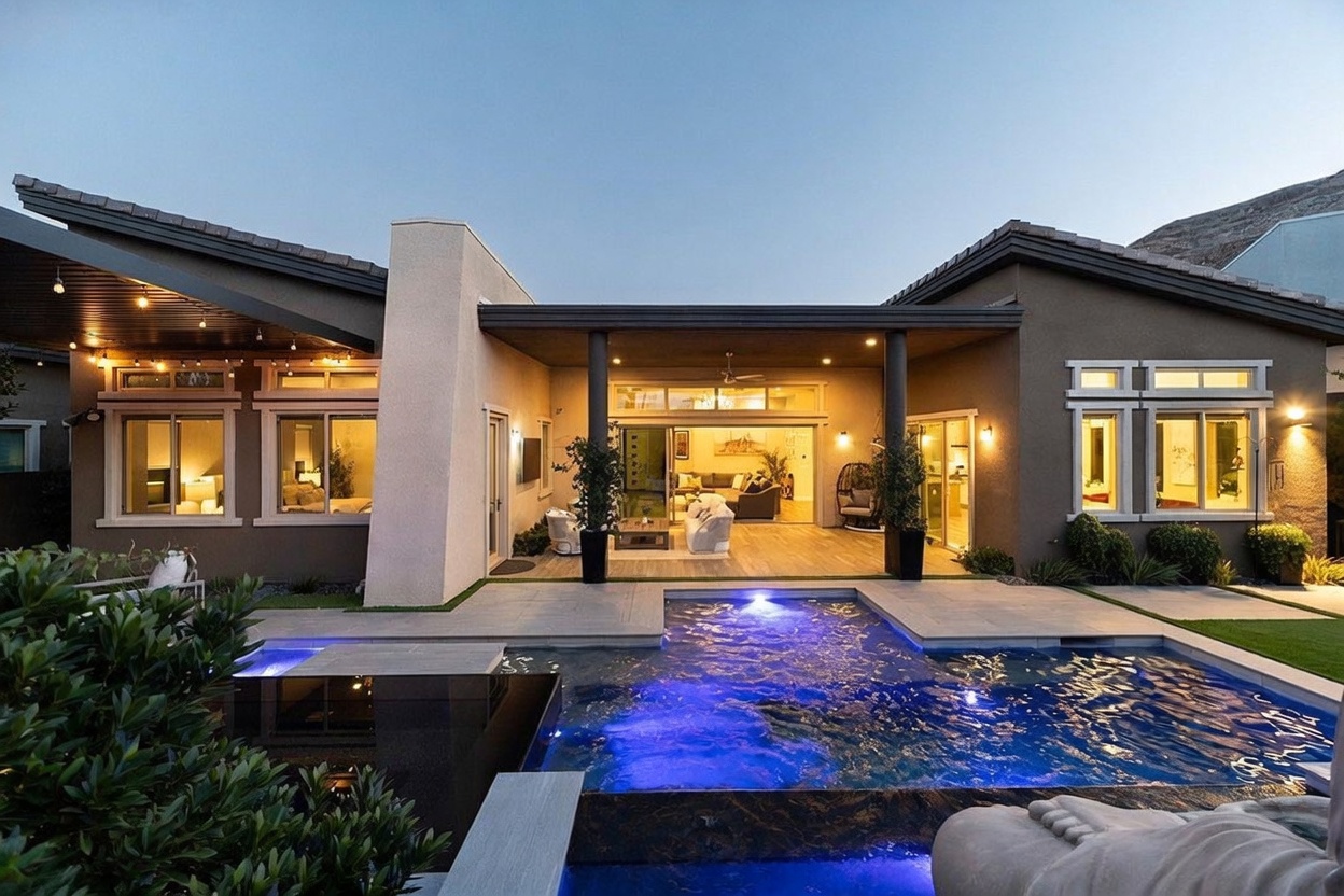 This rear view of a modern residence showcases a seamless transition between indoor and outdoor living spaces, featuring a covered patio that opens into a bright living area. A custom-designed swimming pool with integrated lighting serves as the focal point of the backyard, complemented by manicured landscaping and a tall, prominent chimney structure. The architectural style is contemporary, emphasizing clean lines, neutral tones, and an inviting atmosphere for entertaining.