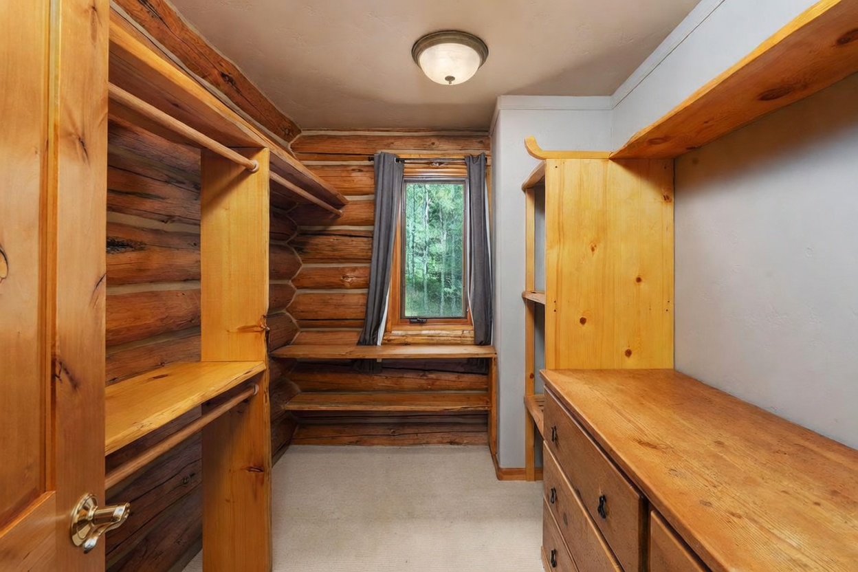 This walk-in closet features rustic log walls and custom light wood shelving units that provide ample storage space. A central window with dark curtains allows natural light to filter in, while a light-colored carpet covers the floor. The room is illuminated by a simple flush-mount ceiling light, creating a warm and cozy atmosphere.