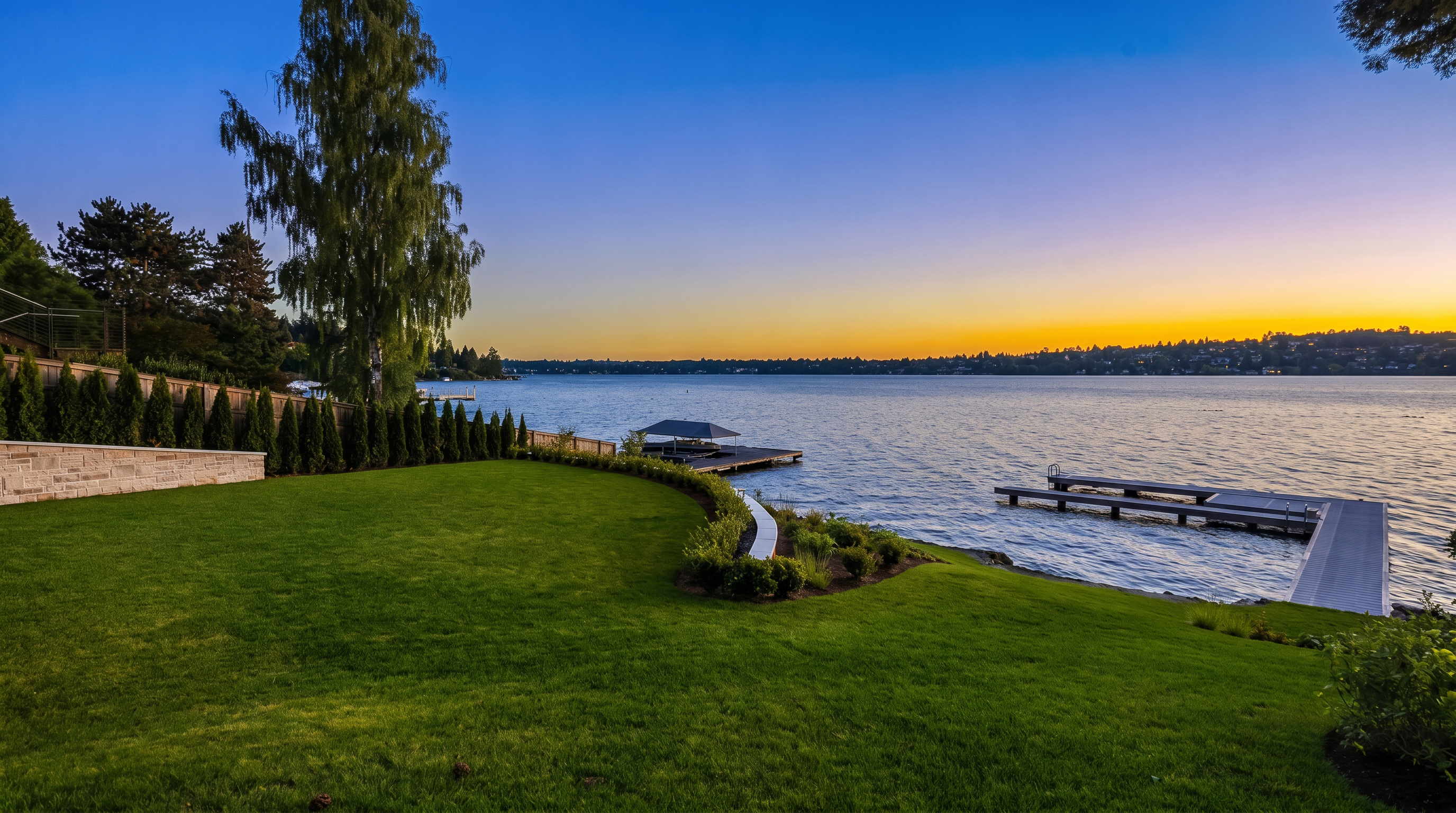 This expansive waterfront backyard features a meticulously manicured lawn that curves gracefully toward the shoreline. A private dock extends into the calm lake, complemented by a smaller covered boat slip, while a stone retaining wall and lush landscaping define the property's edge. The scene is captured at sunset, with a vibrant orange and blue sky reflecting over the water, creating a serene and luxurious atmosphere.