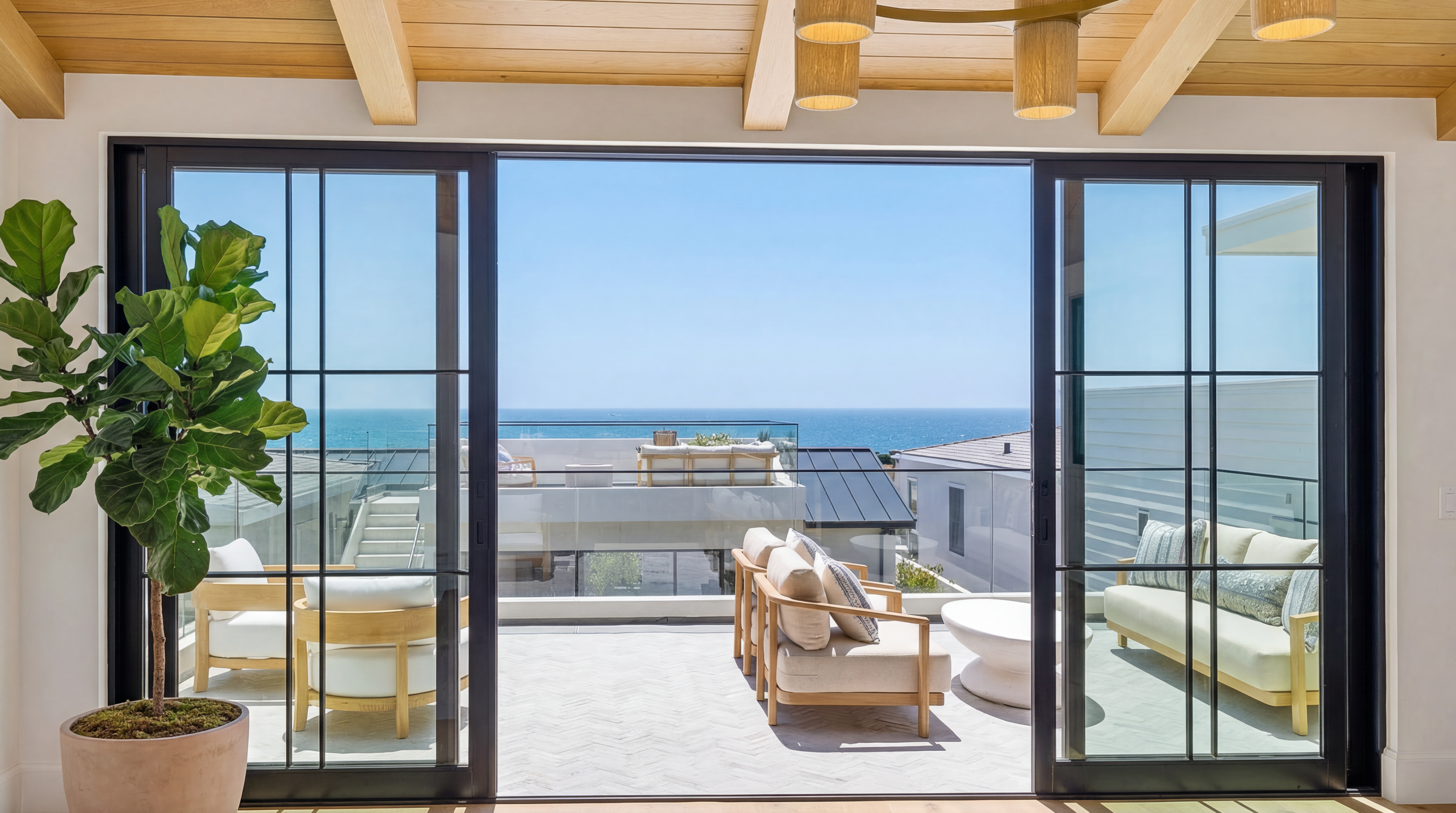 This living room is characterized by large floor-to-ceiling sliding glass doors that open onto a spacious sun-drenched balcony with stunning ocean views. The interior features warm wood-paneled ceilings, a large potted fiddle leaf fig, and a seamless transition to the outdoor seating area. The space exudes a luxury coastal aesthetic, perfect for relaxing and enjoying the expansive horizon.
