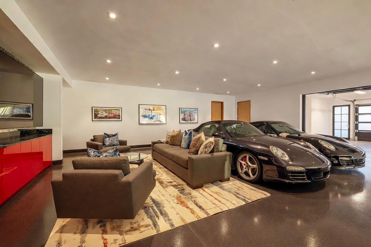 This unique, high-end garage space doubles as a sophisticated lounge area, featuring a plush sofa and armchairs arranged on a patterned area rug. Two luxury sports cars are parked directly adjacent to the seating area, highlighting the space's dual-purpose design. The room is finished with sleek, dark flooring and recessed lighting, creating a polished and modern atmosphere.