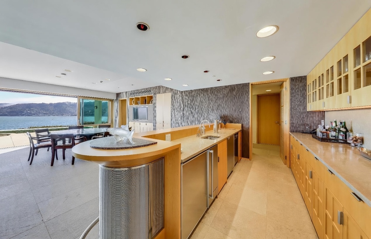 This modern kitchen features light wood cabinetry, a long stone-topped island with a built-in sink, and stainless steel appliances. The space opens directly to a dining area with large glass sliding doors that provide an expansive view of the water and mountains beyond. The room is illuminated by recessed lighting and maintains a sleek, open-concept design.