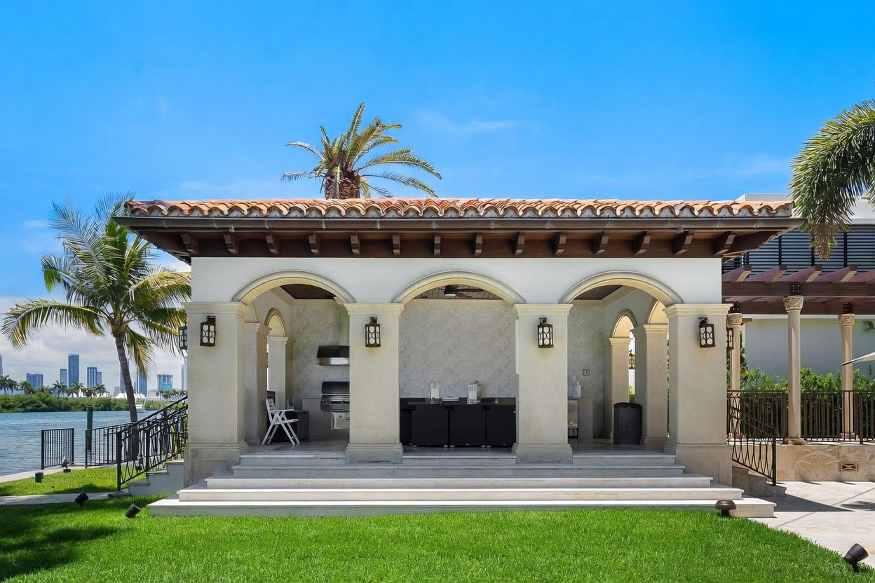 This image showcases a luxurious, Mediterranean-style outdoor pavilion or summer kitchen overlooking a waterfront. The structure features elegant arched openings, stone columns, and a terracotta tile roof, housing a built-in grill and bar area. The scene is set against a lush green lawn with a scenic view of a distant city skyline across the water, creating an inviting and sophisticated atmosphere for outdoor entertaining.