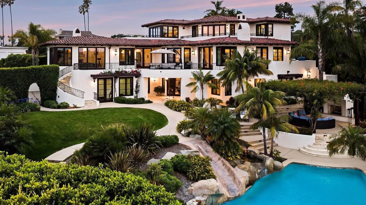 This expansive Mediterranean-style estate features a multi-level rear facade with white stucco walls, dark wood-framed windows, and a classic red tile roof. The property boasts a lush backyard with a curved lawn, a stone-accented water feature with a slide leading into a pool, and a shaded outdoor seating area with an integrated fireplace. The perspective is captured from an elevated angle, showcasing the seamless integration of luxury indoor-outdoor living spaces.