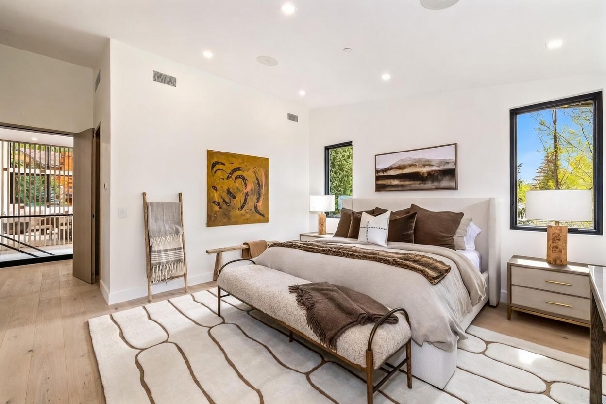 This primary bedroom features a neutral color palette with warm wood flooring and a large, patterned area rug. A plush bed with layered brown and white bedding serves as the focal point, complemented by a textured bench at the foot and matching bedside lamps. The room is brightened by large windows that offer views of the outdoors, while a piece of abstract art adds a bold touch to the minimalist wall decor.