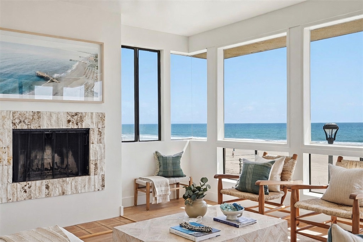 This bright living room features a prominent stone-clad chimney breast and expansive windows that provide panoramic views of the ocean and beach. The space is furnished with light wood chairs, a patterned coffee table, and a bench seat, creating a relaxed, coastal-inspired atmosphere. The neutral color palette and natural light emphasize the seamless connection between the indoor living area and the outdoor scenery.