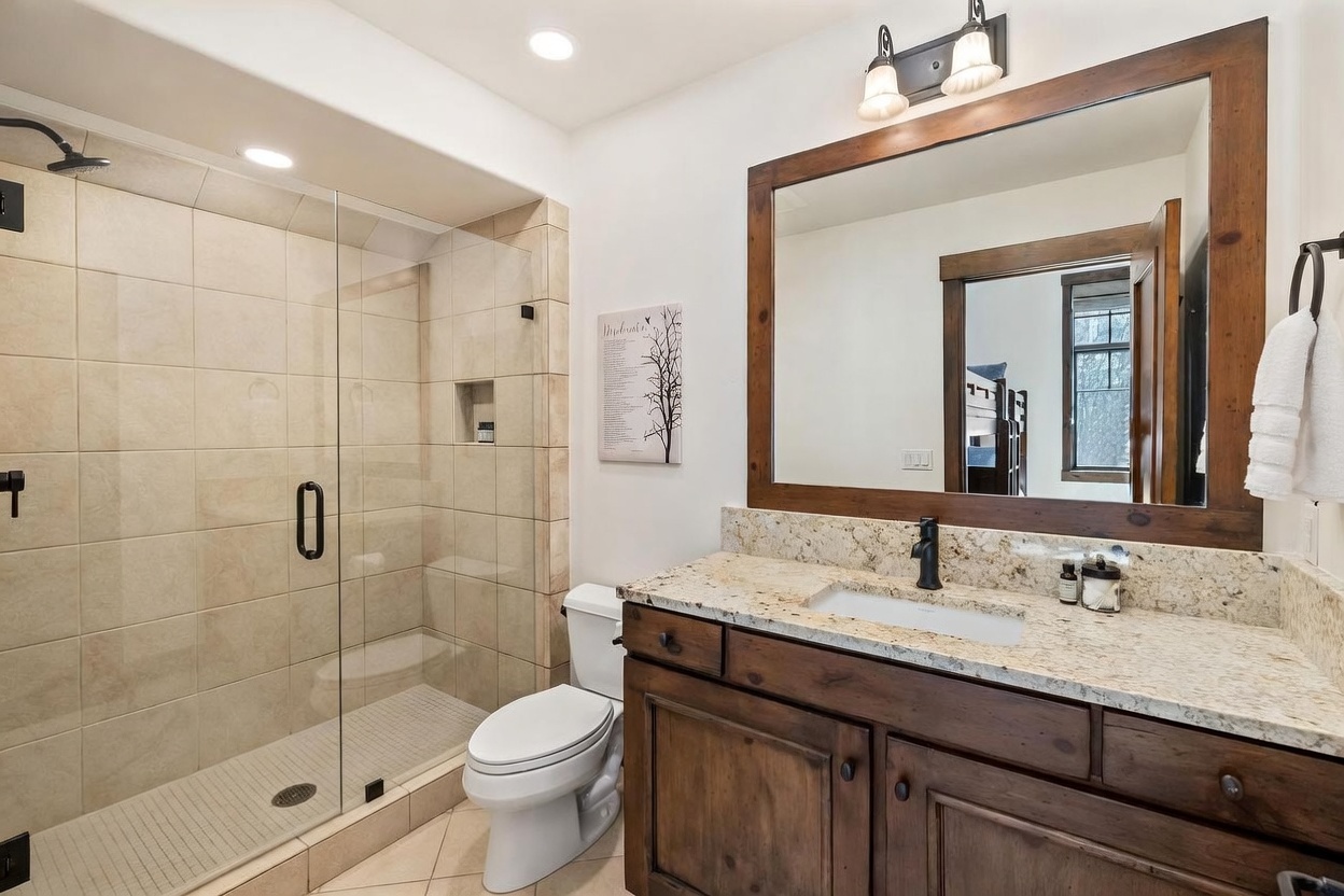 This bathroom features a rustic aesthetic with a dark wood vanity topped by a light-colored stone countertop and a framed mirror. To the left, a walk-in shower is enclosed by a glass panel and finished with neutral-toned wall tiles. The space is completed by a white toilet and warm lighting, creating a cozy and functional atmosphere.