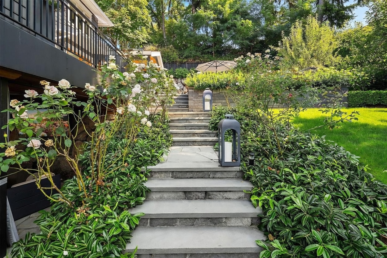 This image captures a beautifully landscaped stone staircase leading up through a lush garden toward a raised deck area. The path is flanked by vibrant green foliage and blooming white roses, with decorative lanterns placed along the steps to provide a warm, inviting atmosphere. The perspective is from the bottom of the stairs, looking upward toward the tiered retaining wall and the shaded outdoor space above.