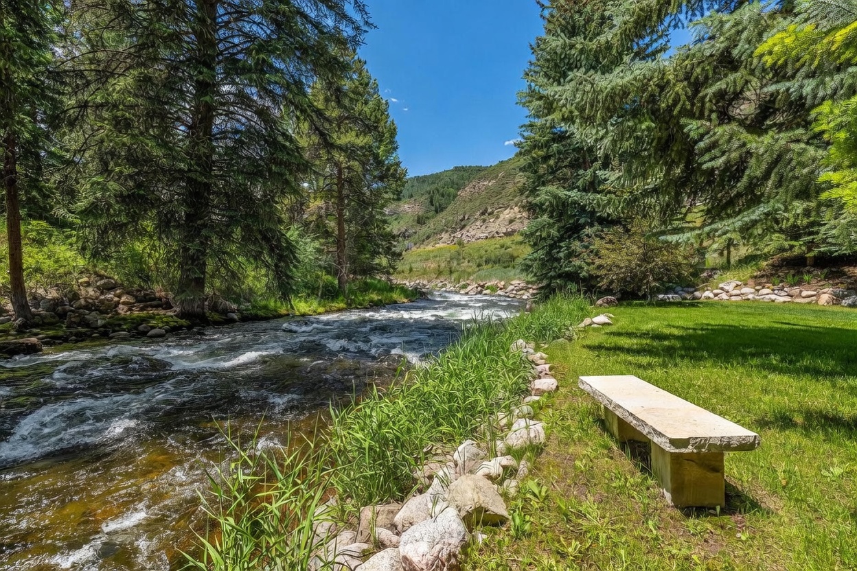 This scenic outdoor space features a stone bench positioned on a manicured lawn overlooking a rushing mountain stream. The landscape is framed by dense evergreen trees and lush greenery, leading the eye toward a rugged, sunlit hillside in the distance. The perspective is captured from a low angle, emphasizing the tranquil connection between the private garden and the natural waterway.