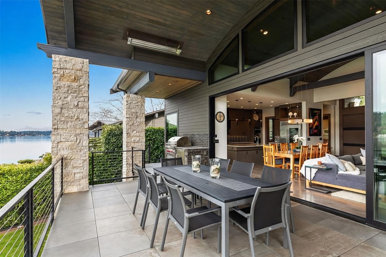 This expansive covered deck offers a seamless transition between indoor and outdoor living through large sliding glass doors. The space features a dark-toned dining set, a built-in outdoor grill station, and stone-clad support columns, all overlooking a serene waterfront view. The modern design emphasizes an open-concept flow, connecting the exterior patio to the interior dining and living areas.