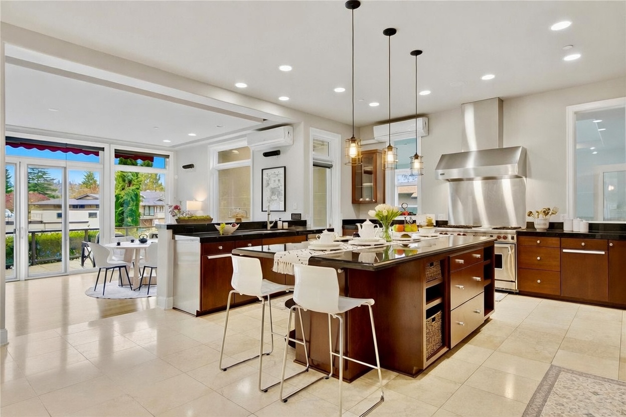 This spacious, open-concept kitchen features a large central island with dark stone countertops and bar seating, illuminated by three pendant lights. The cabinetry is finished in a rich, dark wood tone, complemented by stainless steel appliances and a professional-style range hood. Large glass doors lead to an adjacent dining area and outdoor deck, creating a bright, airy atmosphere with a seamless transition between indoor and outdoor living spaces.