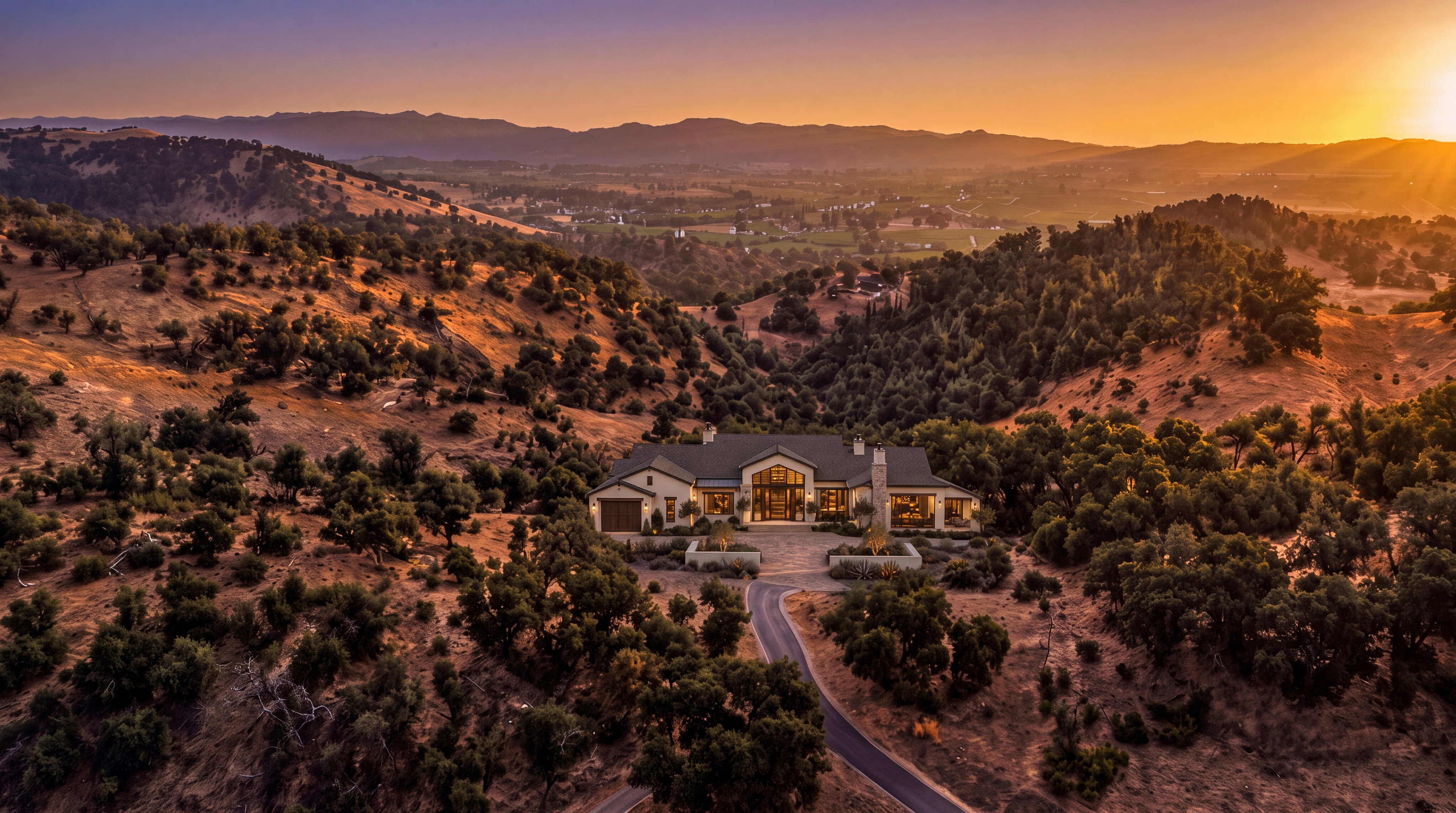 This stunning aerial view captures a sprawling, custom-designed estate nestled within a picturesque, rugged landscape of rolling hills and native trees. The home features a sophisticated architectural style with a grand entrance, warm exterior lighting, and a long driveway leading up to the property, creating a sense of luxury and seclusion. The breathtaking sunset backdrop over the distant mountain range enhances the serene, upscale atmosphere of this unique hilltop residence.