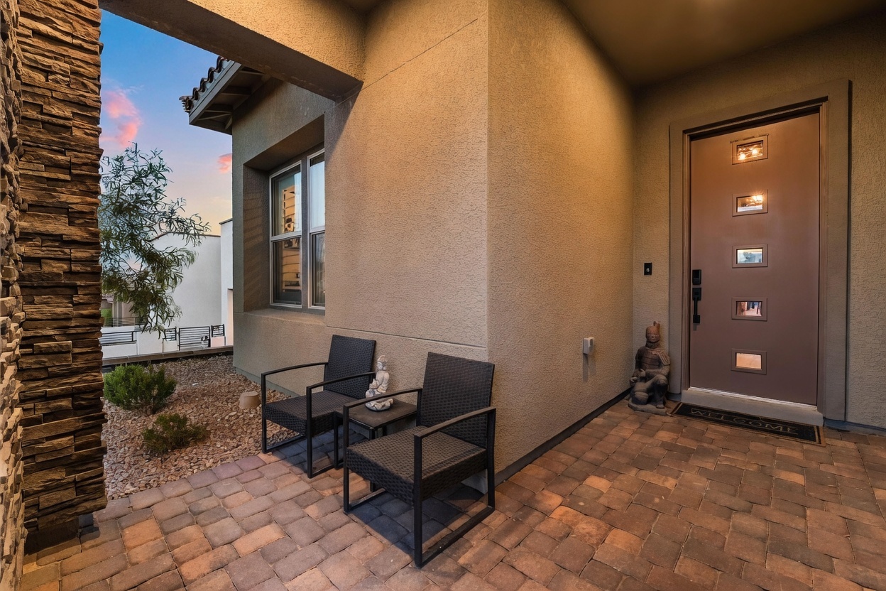 This inviting front entryway features a paved patio area with two dark wicker chairs and a small side table, creating a cozy seating nook. The entrance is defined by a modern brown door with rectangular glass inserts and a stone-clad pillar to the left, set against a textured stucco exterior. A small decorative statue sits near the door, adding a personalized touch to this well-maintained and welcoming home entrance.