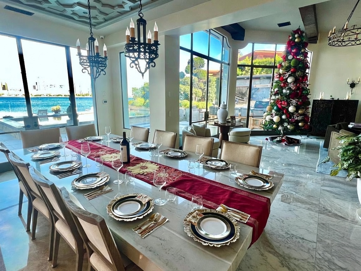 This elegant dining room features a long, stone-topped table set for a formal meal, surrounded by upholstered chairs and illuminated by ornate iron chandeliers. Large windows provide expansive views of the water and outdoor patio, while a decorated Christmas tree adds a festive touch to the corner of the room. The space is characterized by its open, airy layout and polished stone flooring, creating a sophisticated atmosphere for entertaining.