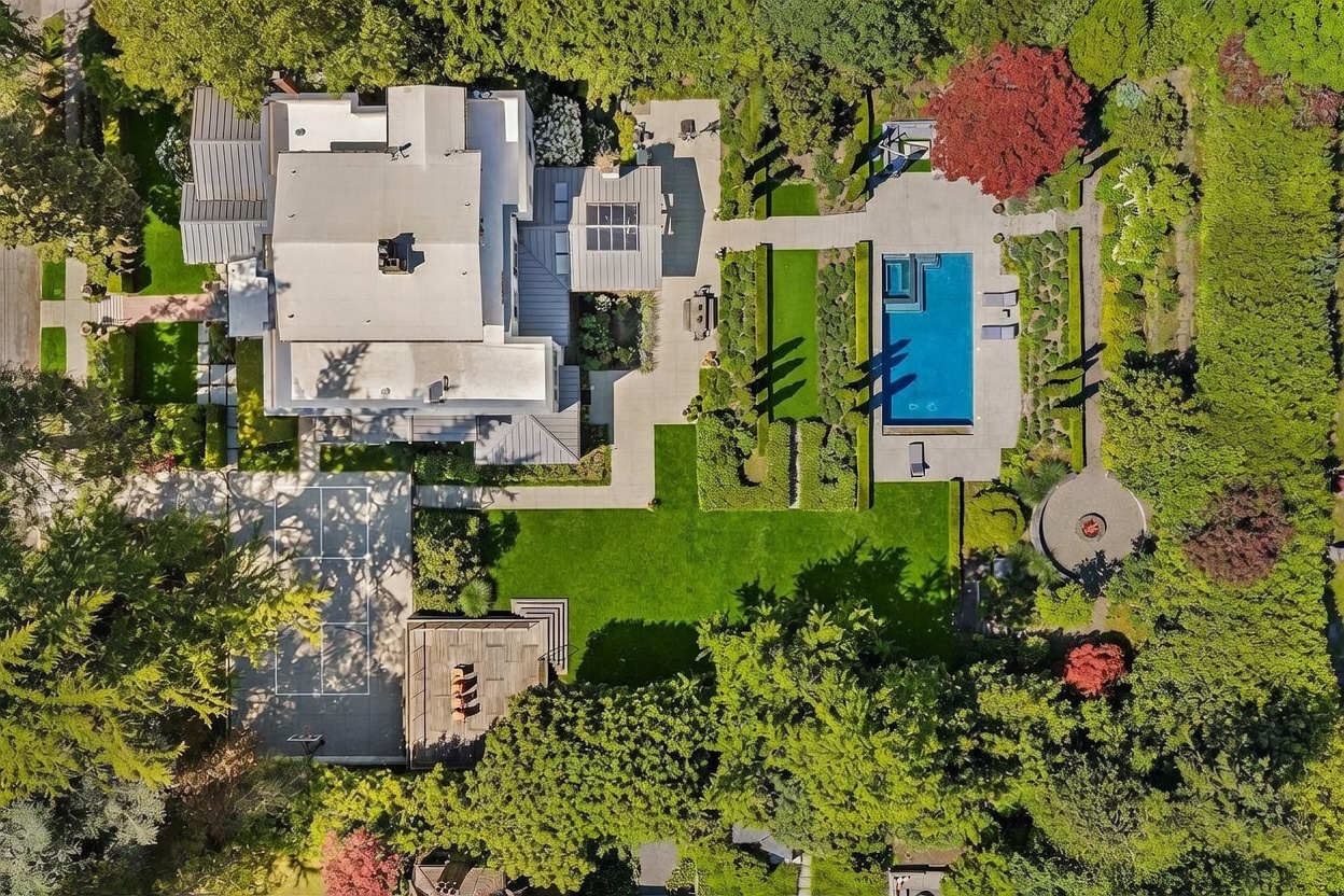 This high-angle aerial view captures a sprawling luxury estate set within a lush, tree-lined landscape. The property features a large modern residence, a rectangular swimming pool, a private sports court, and meticulously manicured gardens with stone pathways. The expansive grounds offer a blend of recreational spaces and serene outdoor living areas, emphasizing privacy and high-end design.
