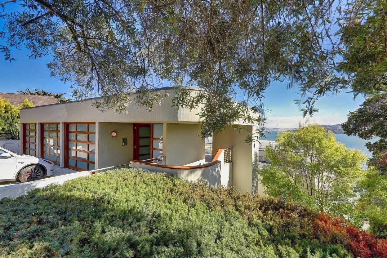 This contemporary home features a unique curved architectural design with a prominent entryway and a multi-car garage with glass-paneled doors. The property is nestled into a hillside, surrounded by lush greenery, and offers a stunning, elevated view of the Golden Gate Bridge in the distance. A white vehicle is parked in the driveway, and a wooden handrail leads toward the entrance, emphasizing the home's integration with its scenic coastal environment.