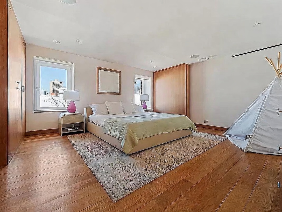 This spacious primary bedroom features warm wood flooring and light-toned walls, creating a bright and airy atmosphere. The room is furnished with a central bed flanked by two nightstands with pink lamps, and includes built-in wood cabinetry along the walls for ample storage. A decorative play tent sits in the corner, adding a playful touch to the otherwise sophisticated and clean living space.