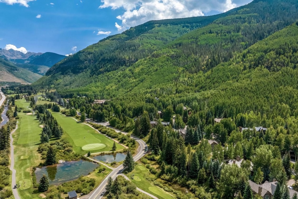 This high-angle aerial view captures a sprawling mountain landscape featuring a lush green golf course winding through a dense forest of pine and aspen trees. A winding road cuts through the property, connecting several secluded residential homes nestled among the trees, with a small pond visible near the foreground. The scene is set against a backdrop of majestic, rugged mountain peaks under a bright, clear blue sky, emphasizing the serene and expansive nature of the location.