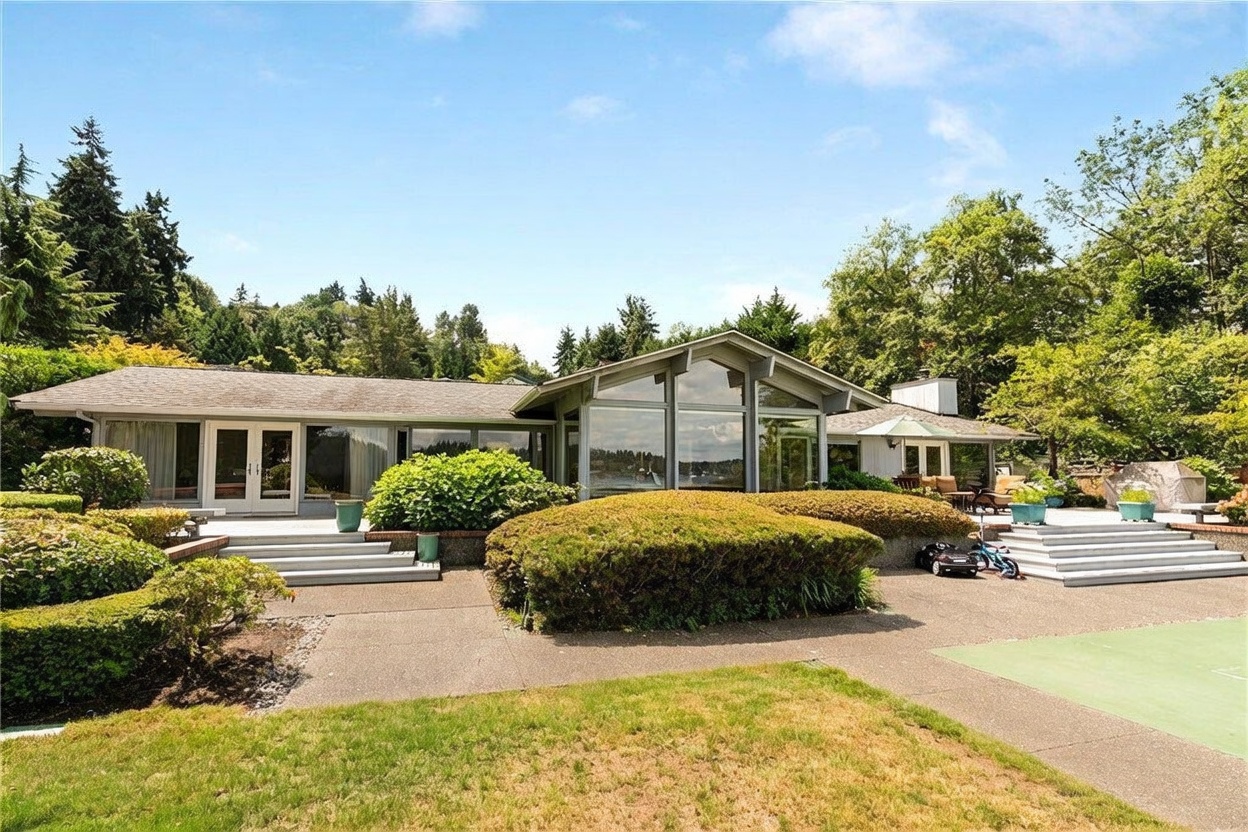 This mid-century modern home features a sprawling, low-profile design with extensive glass walls that offer a seamless connection to the outdoors. The rear exterior showcases a multi-level concrete patio, manicured landscaping with rounded shrubs, and a wide, open lawn area. The architectural style emphasizes horizontal lines and natural light, creating an inviting and serene atmosphere.