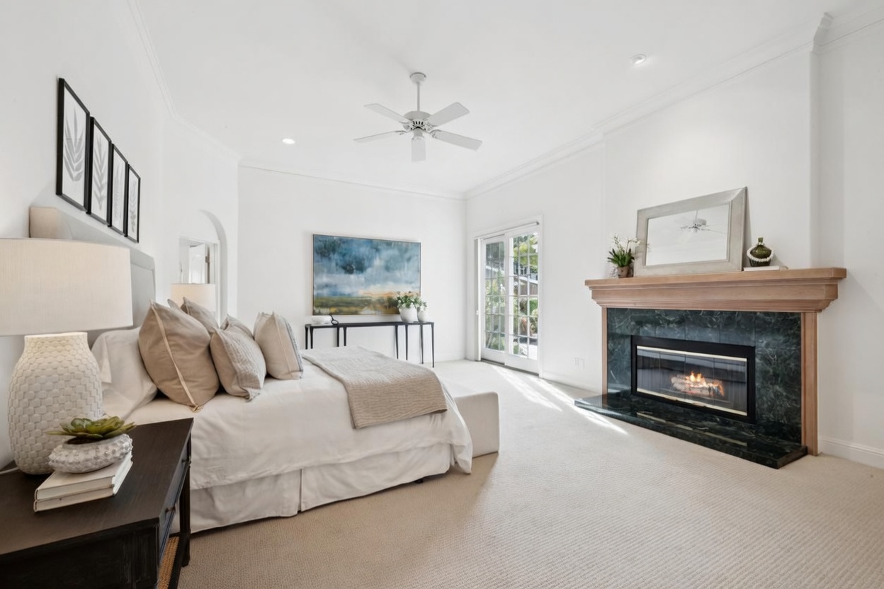 This elegant primary bedroom features a cozy fireplace with a dark green marble surround and wood mantel, creating a sophisticated focal point. The room is neutrally toned with soft beige carpeting, a large plush bed, and crisp white walls that enhance the spacious and tranquil atmosphere. Natural light floods the room through glass doors, highlighting the clean, modern aesthetic and thoughtful decor.