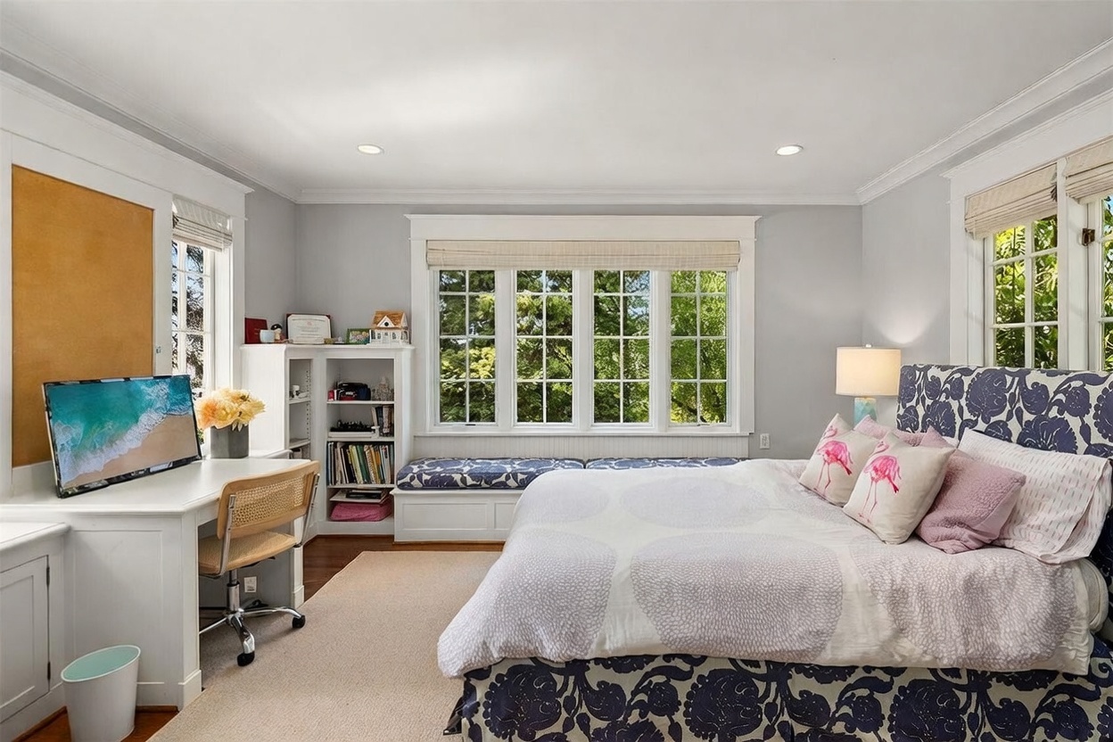 This bright and inviting bedroom features a large bed with a bold blue and white floral patterned headboard and matching bedding. A built-in desk and shelving unit are positioned against the wall, creating a functional workspace, while a window seat with a patterned cushion sits beneath a row of windows. The room is finished with light gray walls, a neutral carpet, and a classic aesthetic that feels both cozy and organized.