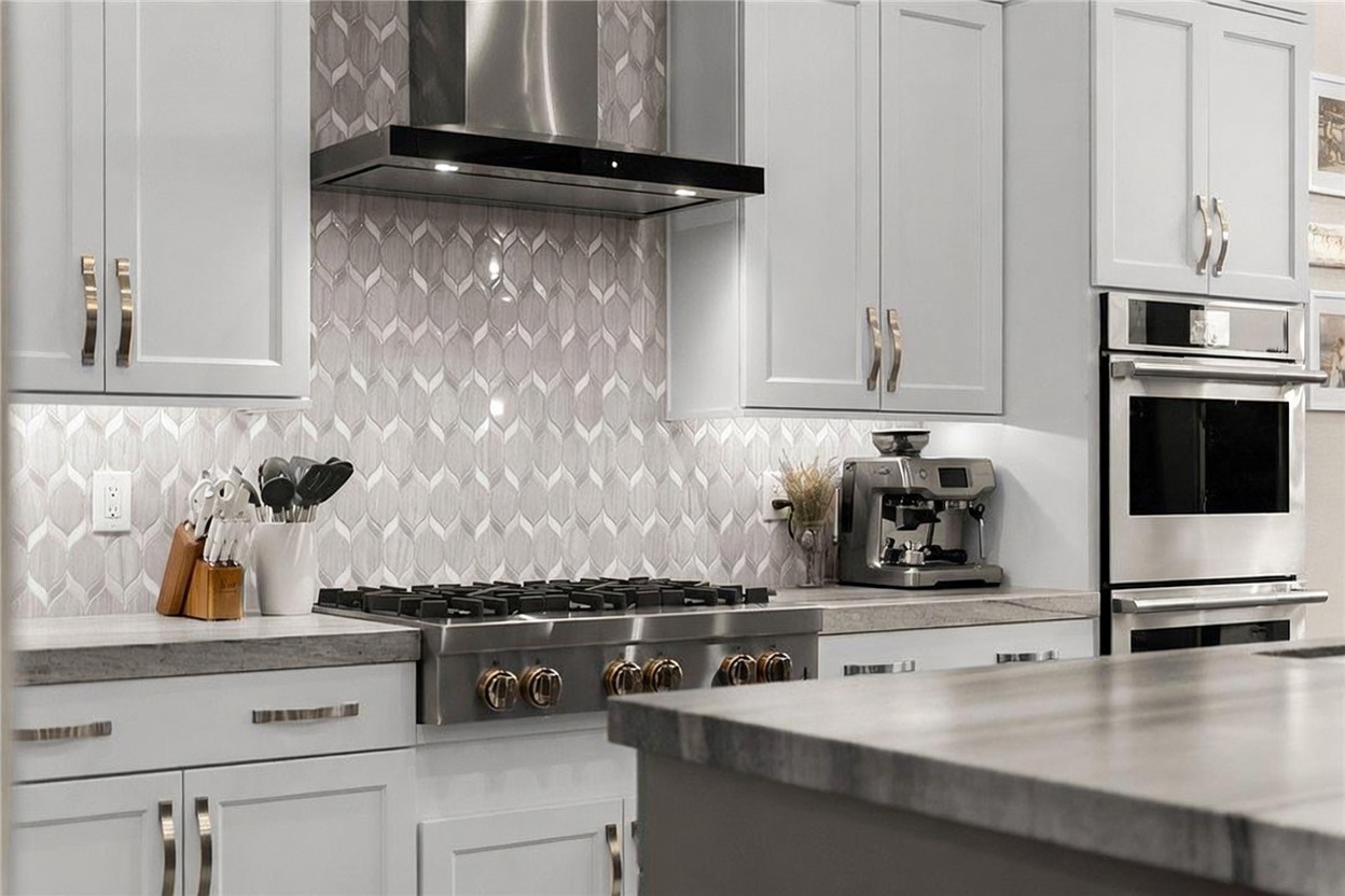 This modern kitchen features crisp white cabinetry paired with a striking patterned tile backsplash and a stainless steel gas range. A professional-style range hood sits above the stove, while a built-in double oven is integrated into the cabinetry on the right. The foreground shows a portion of a kitchen island with a neutral-toned countertop, creating a clean and functional culinary space.