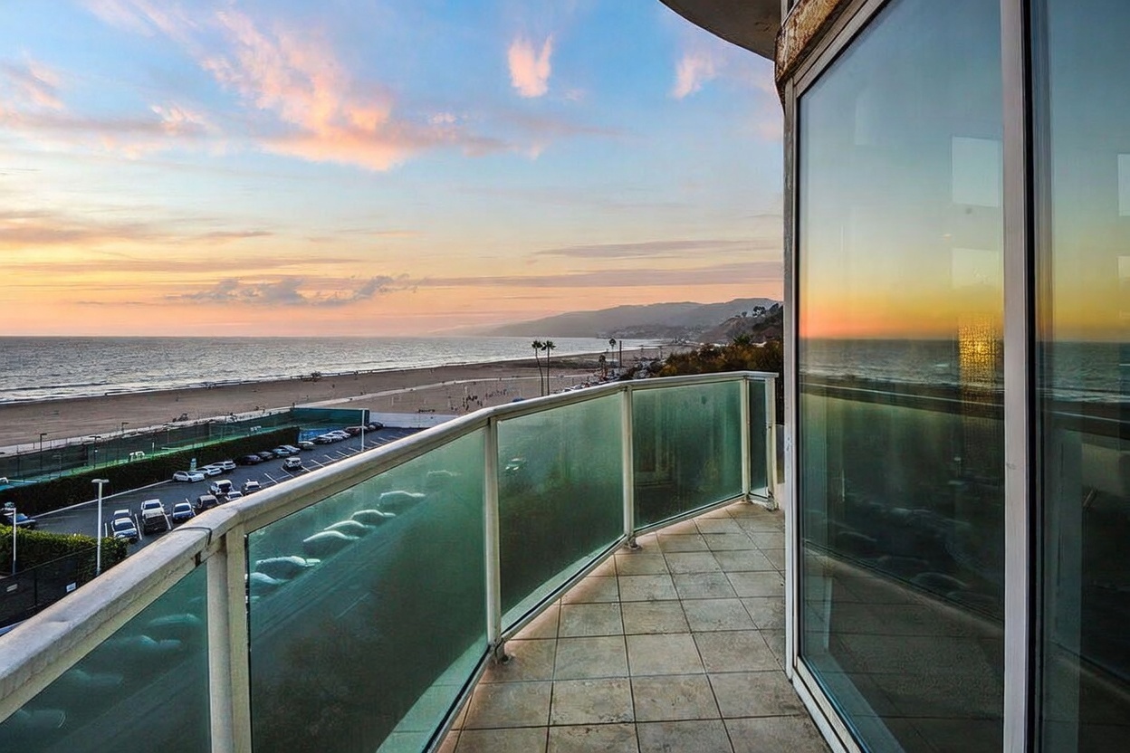This balcony offers a panoramic, elevated view of the coastline, beach, and ocean at sunset. The space features light-colored tile flooring and a glass-paneled railing that ensures an unobstructed view of the horizon. Large glass doors provide seamless indoor-outdoor transition, capturing the warm, golden light of the setting sun.