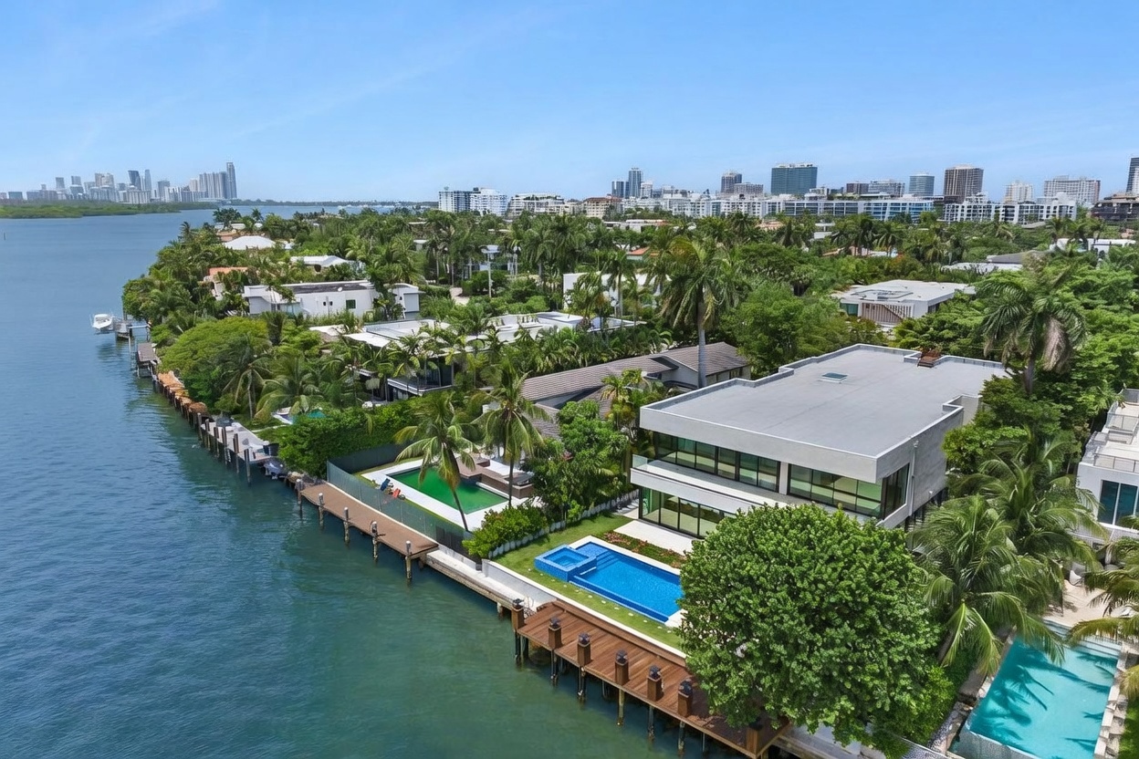 This high-angle aerial shot captures a luxurious modern waterfront estate featuring a sleek, multi-level residence with expansive glass walls. The property includes a private swimming pool, a wooden dock extending into the calm bay, and lush tropical landscaping. In the background, a sprawling cityscape stretches along the horizon under a clear blue sky, emphasizing the prime coastal location.
