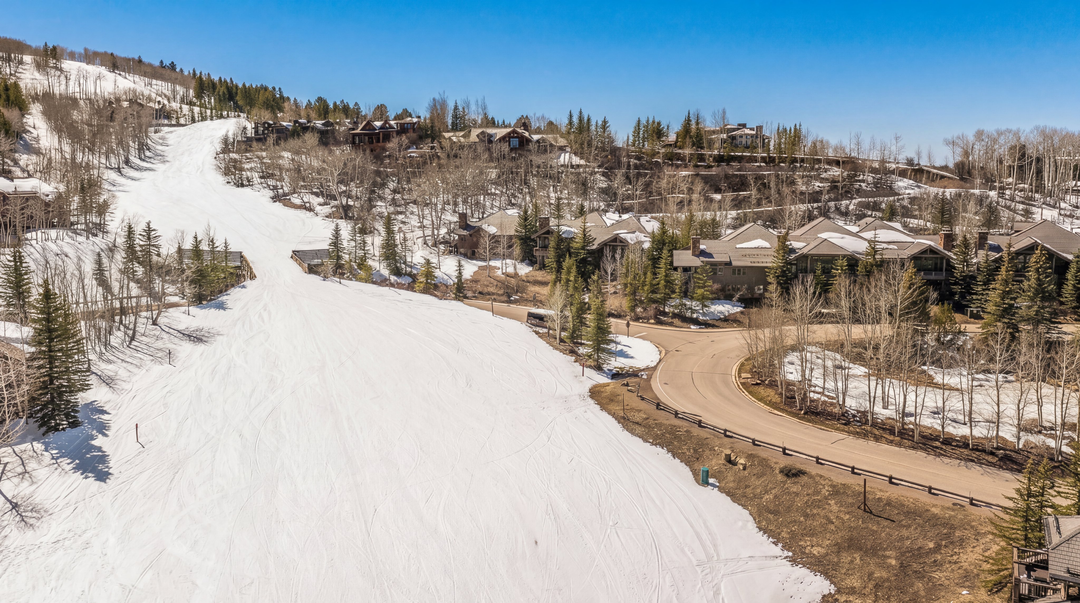 This stunning aerial view captures a luxury residential community nestled along the snow-covered slopes of a mountain resort. The scene features upscale multi-story homes integrated into the natural landscape, alongside a winding access road that provides scenic connectivity. The contrast between the expansive, groomed ski run and the sophisticated wooden architecture highlights the property's prime location in a premier winter destination.