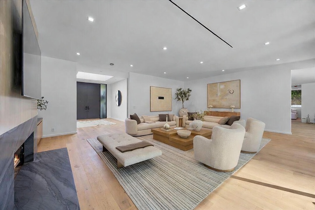 This spacious, open-concept living room features light wood flooring and a neutral color palette, creating an airy and sophisticated atmosphere. The room is anchored by a large, textured area rug, a low-profile bench, and a grouping of plush, cream-colored seating arranged around a tiered wooden coffee table. A tall, dark stone-clad chimney breast stands to the left, while minimalist artwork and a potted plant add subtle decorative touches to the bright, recessed-lit space.