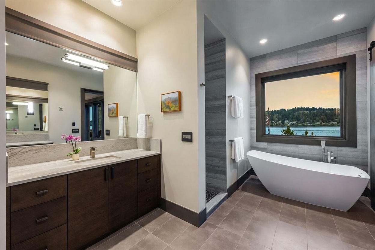 This primary bathroom features a sleek, modern design with a dark wood vanity topped with a light-toned stone counter and a large mirror. To the right, a freestanding soaking tub is positioned beneath a large window that offers a scenic view of the water and surrounding trees. The space is finished with neutral-toned floor tiles and a walk-in shower area with textured wall tiles, creating a serene and luxurious atmosphere.