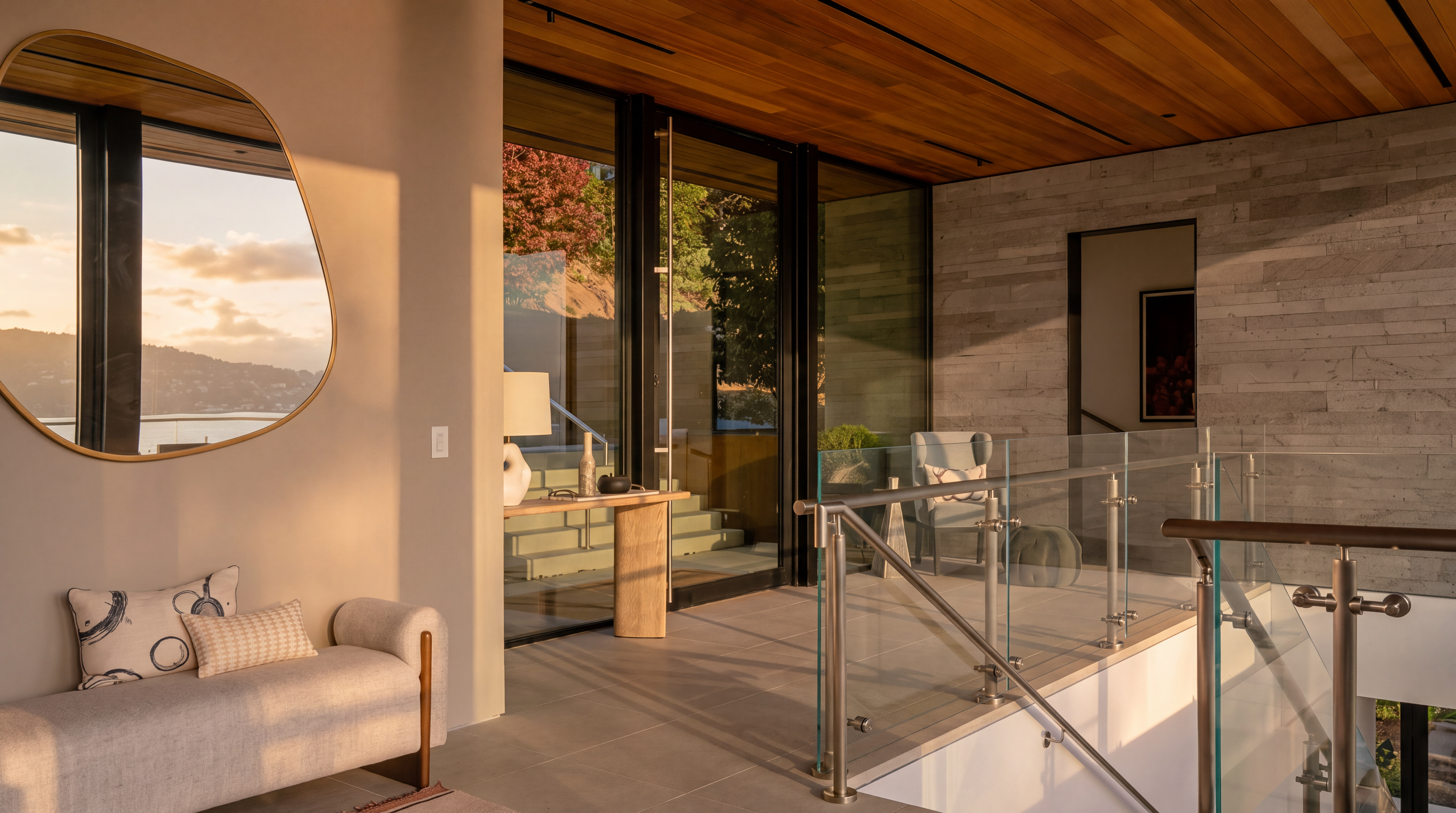This sophisticated interior space features a landing area with a light-toned bench and a unique, organic-shaped mirror reflecting a scenic outdoor view. The area transitions into a staircase with a glass railing and metal hardware, set against a textured stone wall and a warm wood-paneled ceiling. Large glass doors provide a seamless connection to the outdoors, creating an airy and modern atmosphere.