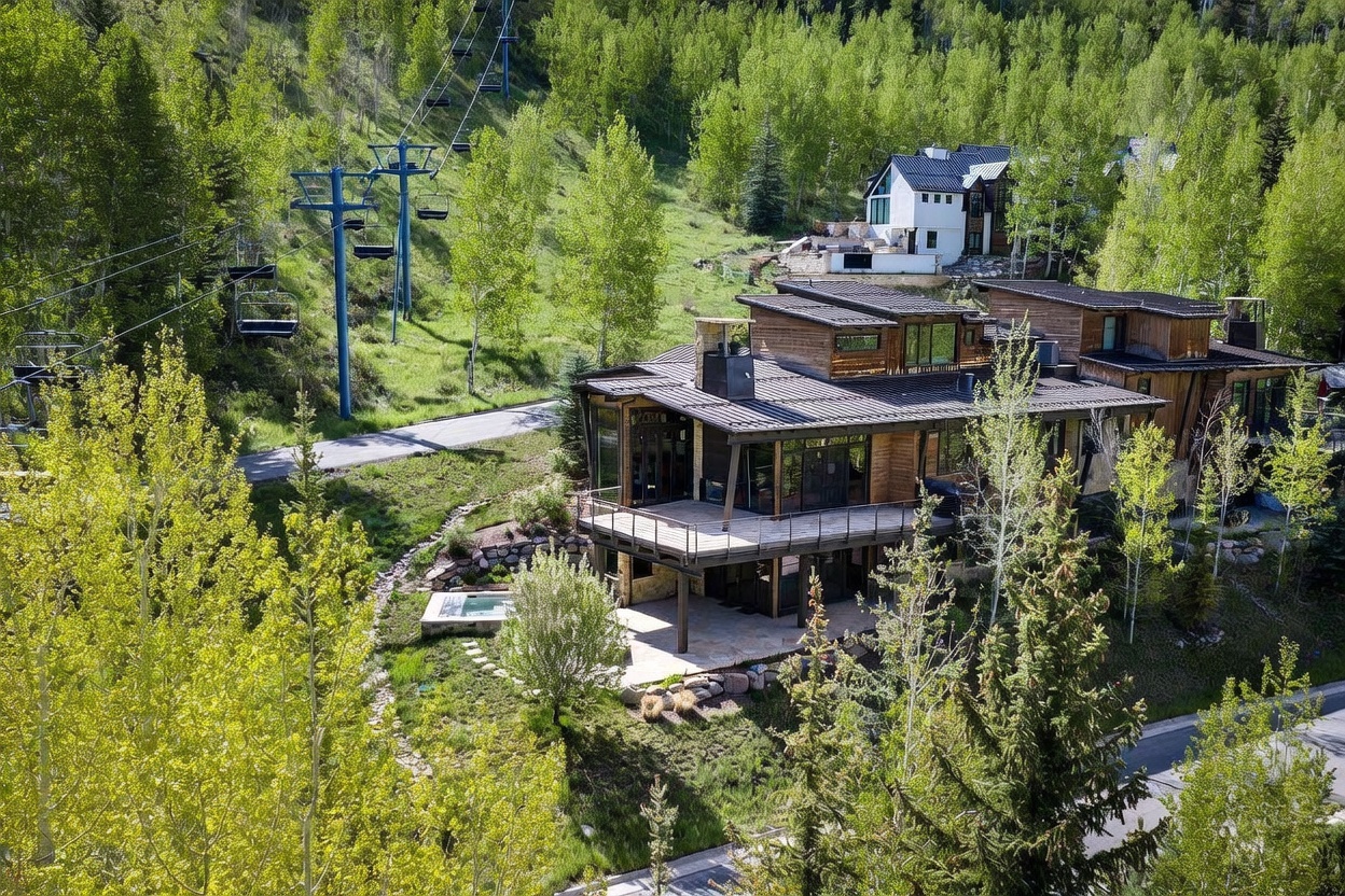 This elevated aerial perspective showcases a contemporary mountain residence nestled into a lush, tree-covered hillside. The home features a multi-level design with expansive wooden decks, a stone-clad chimney, and a private ground-level spa area, all set against a backdrop of vibrant green foliage and a ski lift. The architectural style blends modern lines with rustic materials, creating a seamless connection to the surrounding natural landscape.