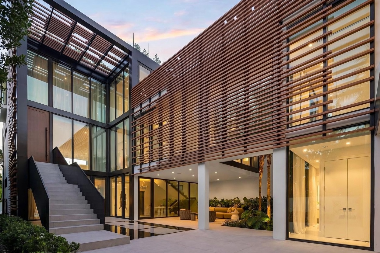 This modern architectural residence features a striking combination of dark metal framing, expansive glass walls, and horizontal wood slat screening. A wide concrete staircase leads to an elevated entryway, while the ground level opens into a covered patio area furnished with outdoor seating. The design emphasizes clean lines and a seamless transition between indoor and outdoor living spaces, complemented by integrated landscape lighting and a small water feature.