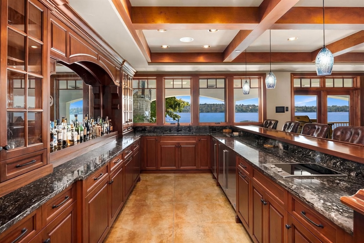 This elegant home bar area features rich, dark wood cabinetry, polished granite countertops, and a stunning view of the lake through large windows. The space is accented by coffered ceilings with recessed lighting and stylish pendant lights hanging over the bar seating area. The warm, inviting atmosphere is enhanced by the natural light and the sophisticated, classic design aesthetic.