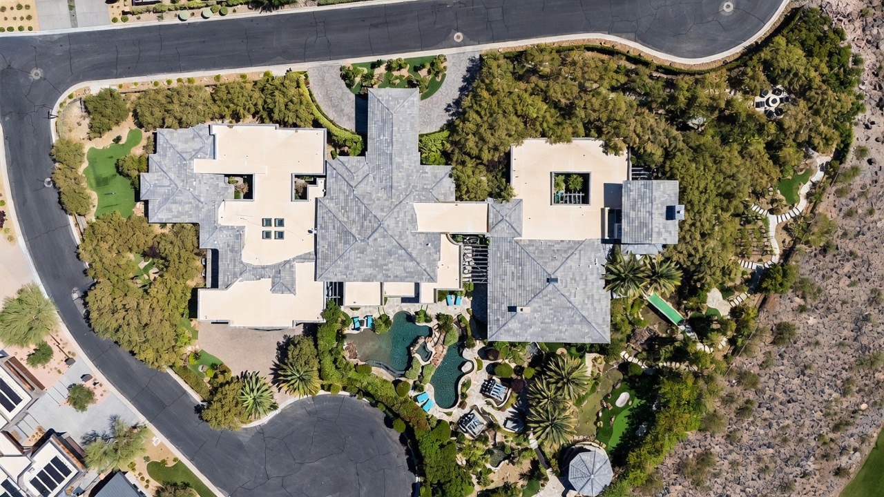 This high-altitude aerial view captures a sprawling, multi-winged luxury estate nestled within a desert landscape. The property features a complex roofline with grey tiles, a central courtyard, and an expansive backyard complete with a custom-shaped swimming pool, lush landscaping, and a private putting green. The home is situated at the end of a cul-de-sac, surrounded by mature trees and rugged terrain, showcasing a private and secluded atmosphere.