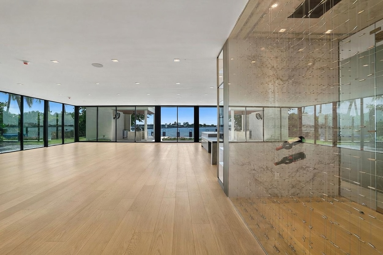 This spacious, open-concept interior features a prominent glass-enclosed wine display on the right, showcasing a modern racking system against a stone wall. The room is characterized by light wood flooring and expansive floor-to-ceiling windows that provide a clear view of the outdoor patio and waterfront beyond. The minimalist design emphasizes clean lines and a bright, airy atmosphere, creating a seamless transition between the indoor living space and the exterior environment.