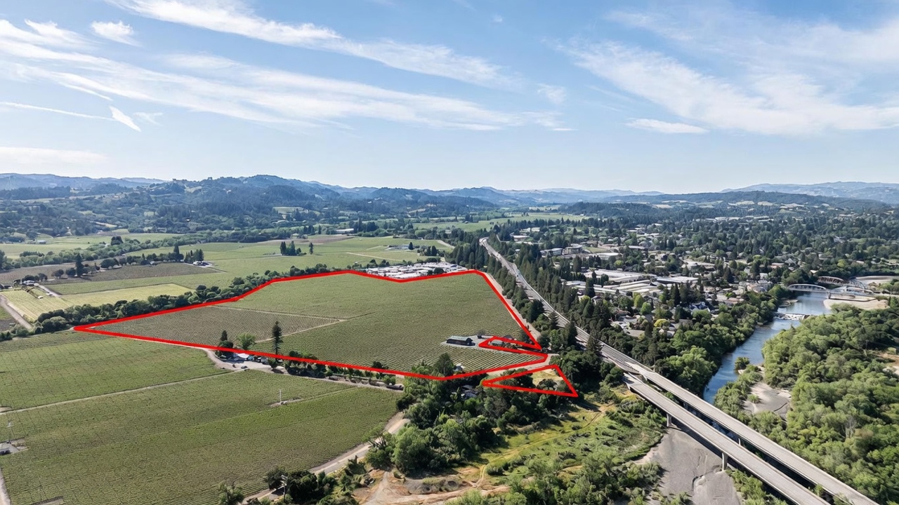 This high-altitude aerial view captures a sprawling agricultural property highlighted by a red boundary outline. The landscape features extensive vineyard rows, a small cluster of buildings, and proximity to a river and a major highway bridge. The perspective provides a comprehensive overview of the land's scale and its strategic location within the surrounding valley.