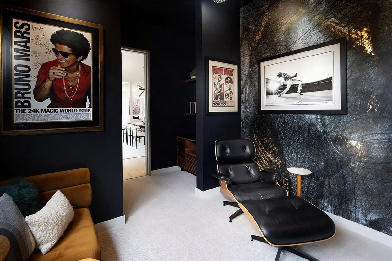 This stylish, music-themed lounge or study features bold black walls and a striking accent wall with a dark, textured forest-inspired mural. The space is furnished with a classic black leather lounge chair and ottoman, complemented by a tan sofa and framed Bruno Mars memorabilia. A doorway leads to an adjacent dining area, creating a sophisticated and moody atmosphere perfect for relaxation or creative work.