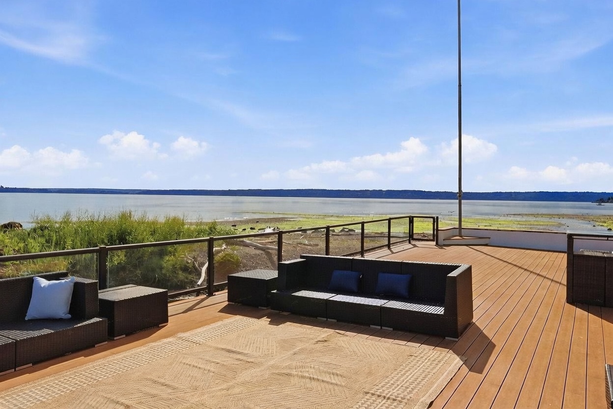 This expansive outdoor deck offers a panoramic view of the water and shoreline, featuring modern dark wicker patio furniture arranged for comfortable seating. The deck is constructed with warm-toned composite planks and is enclosed by a sleek glass and metal railing system that preserves the scenic vista. A large, textured area rug anchors the seating area, creating a sophisticated and inviting space for relaxation or entertaining.