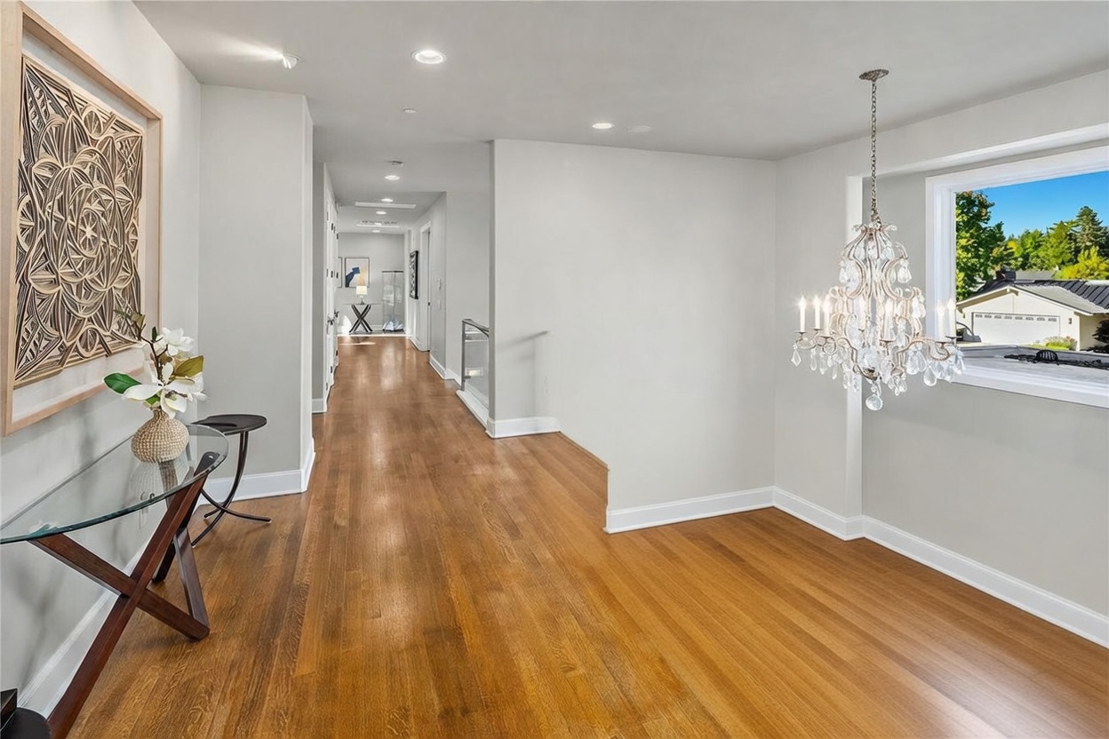 This bright upstairs hallway features warm-toned wood flooring and a sophisticated glass-paneled railing that overlooks the lower level. A large, ornate crystal chandelier hangs prominently, adding a touch of elegance, while a decorative wall art piece and a glass-topped console table provide a welcoming aesthetic. The perspective looks down the length of the hallway, highlighting the clean lines and open, airy feel of the home's upper floor.