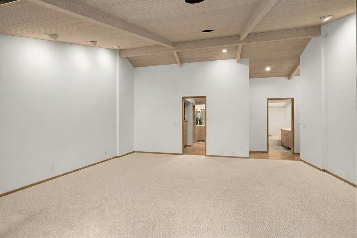 This spacious, open-concept living area features a vaulted ceiling with exposed light-toned wood beams and recessed lighting. The room is finished with neutral-colored carpeting and plain white walls, providing a blank canvas for interior design. Two doorways lead into adjacent rooms, one revealing a glimpse of a bathroom vanity, creating a sense of flow throughout the home.