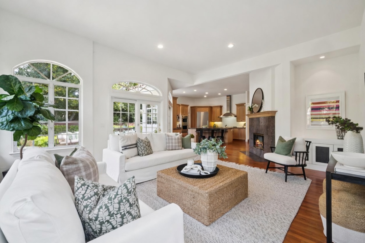 This bright and inviting living room features a plush white sofa, a large textured woven coffee table, and a cozy fireplace with a stone surround. The open-concept space flows seamlessly into a warm wood-toned kitchen, highlighting a well-lit and cohesive interior design. Large arched windows provide ample natural light and a pleasant view of the outdoor greenery.