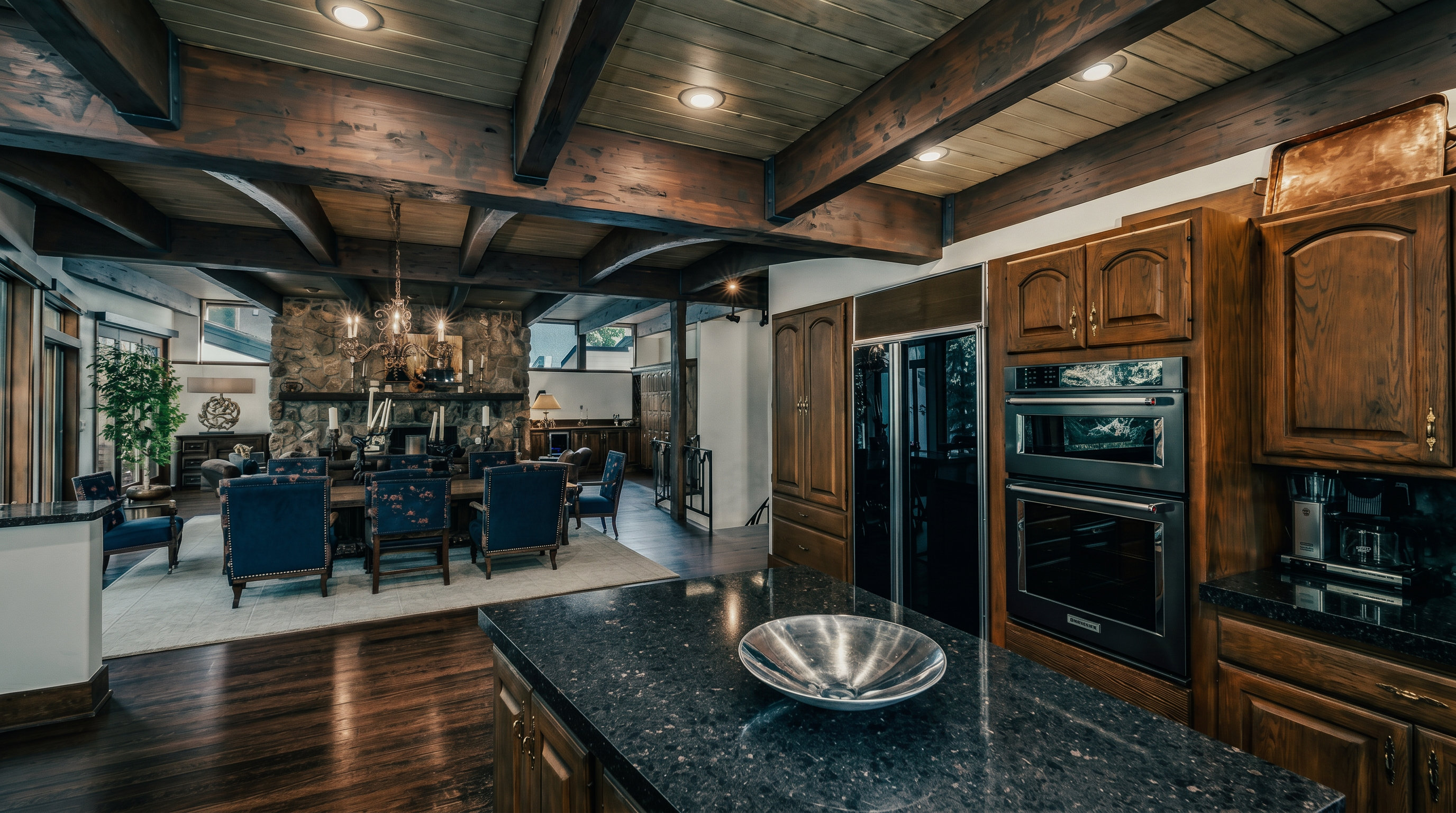 This spacious kitchen features warm-toned wood cabinetry paired with dark, speckled stone countertops and a central island. The room is defined by a striking exposed beam ceiling and recessed lighting, creating a rustic yet refined atmosphere. In the background, a stone-clad chimney breast anchors the adjacent living area, providing a cohesive and open-concept flow throughout the home.