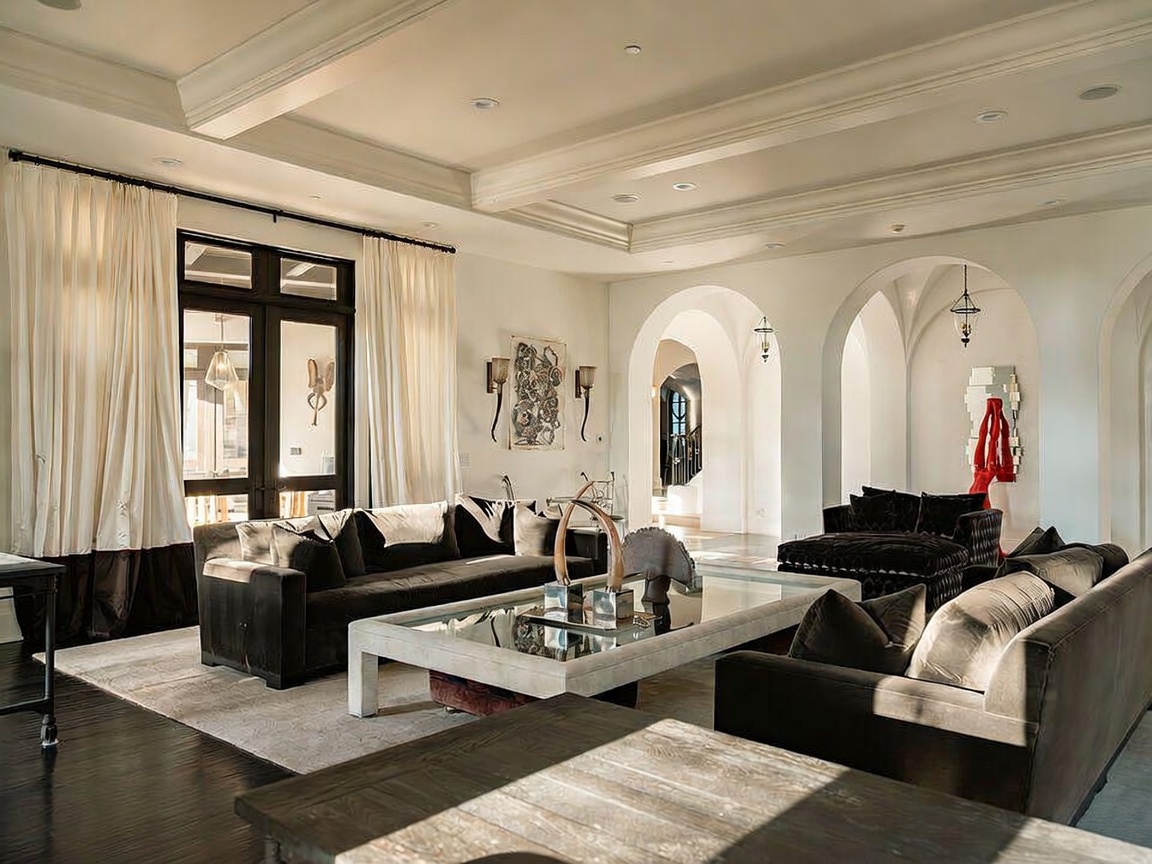This spacious living room features a sophisticated, neutral color palette with dark velvet sofas and a large, light-toned coffee table as the centerpiece. The room is defined by elegant architectural arches that lead to adjacent spaces, complemented by tall windows draped in floor-length curtains. The overall aesthetic is one of refined luxury, enhanced by recessed lighting and a clean, open floor plan.