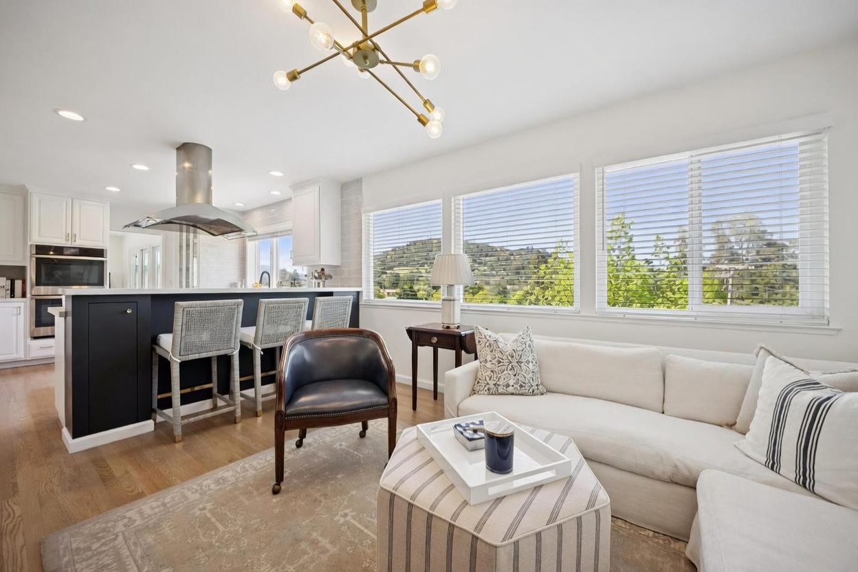 This open-concept living space seamlessly integrates a kitchen and lounge area, featuring a large white sofa, a patterned ottoman, and a dark leather accent chair. The room is bathed in natural light from a row of large windows that offer a scenic view of the surrounding hills. A modern, multi-armed brass chandelier serves as a focal point above the transition between the kitchen island and the living area.