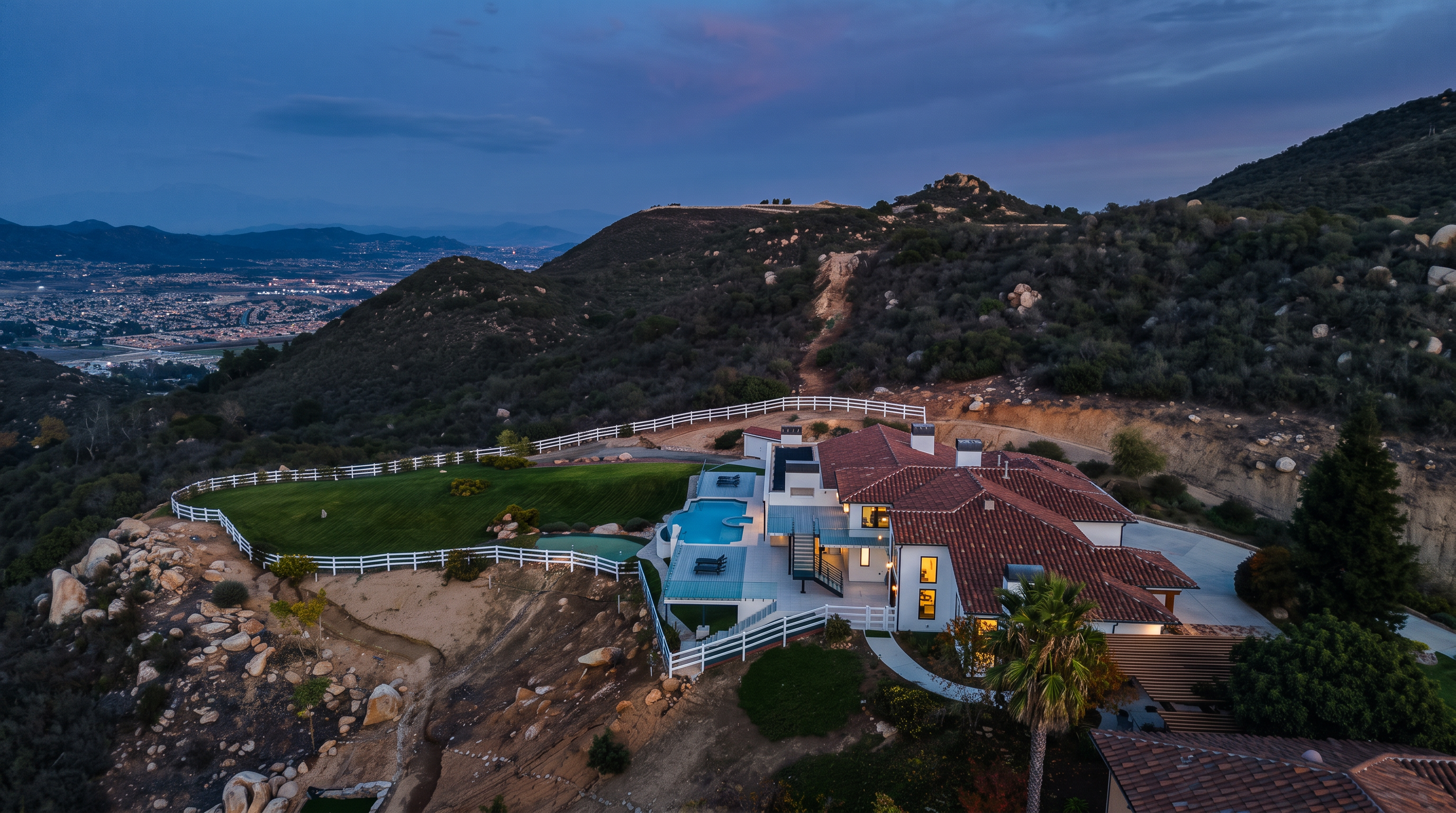 This breathtaking aerial perspective showcases a luxurious hilltop estate nestled into a dramatic landscape. The residence features classic Mediterranean-style architecture with a tile roof, an expansive tiered backyard, a stunning pool and spa area, and sweeping panoramic views of the valley below. The property perfectly blends refined living with the rugged beauty of its natural surroundings.