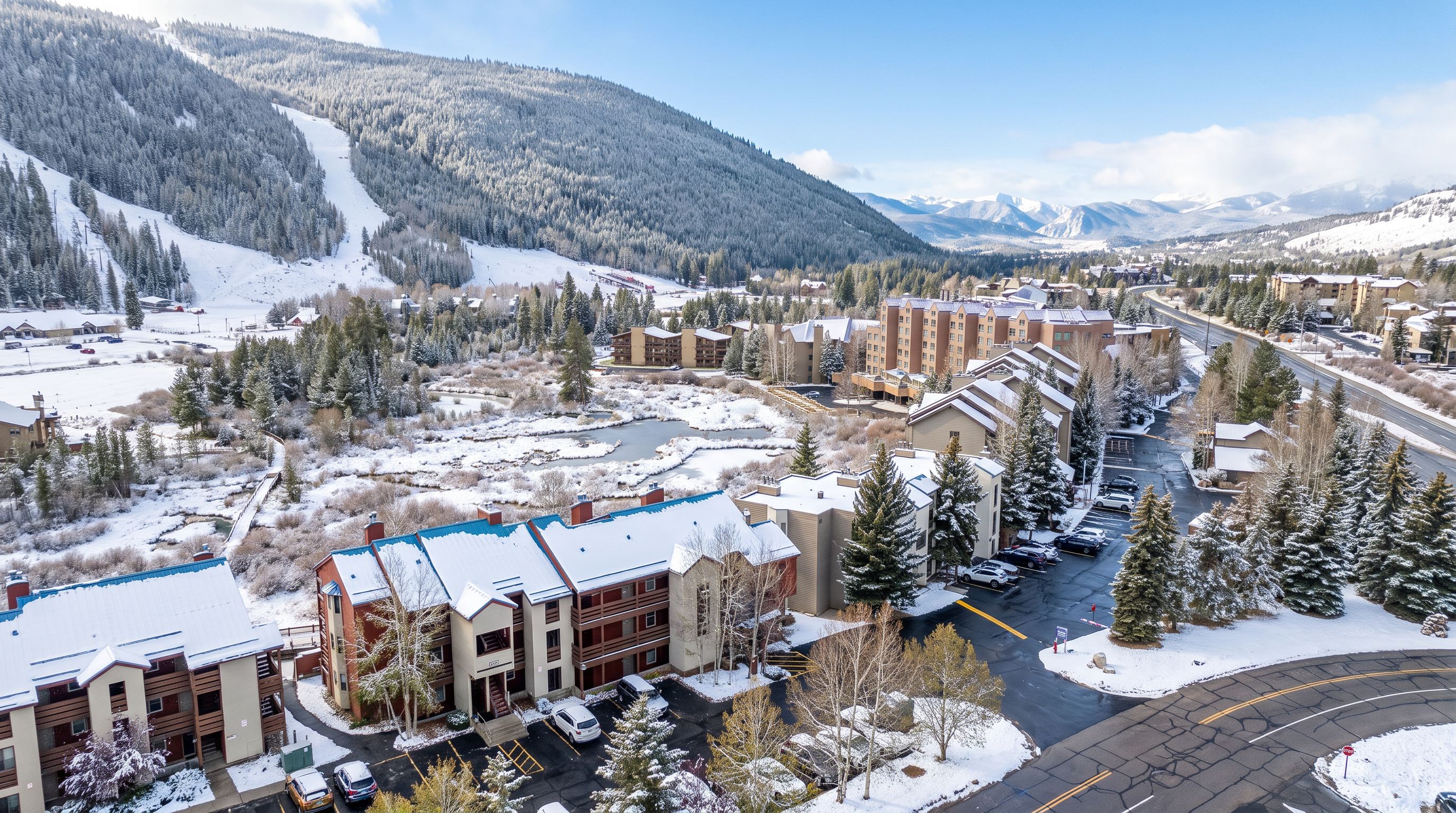 This high-angle aerial perspective captures a multi-building residential complex nestled in a scenic mountain valley. The scene features buildings with distinctive blue metal roofs, ample parking areas, and a lush, marshy wetland area that separates the residential structures from the forested mountain slopes in the background. A winding road runs along the right side of the frame, emphasizing the property's accessible yet tranquil location near nature.